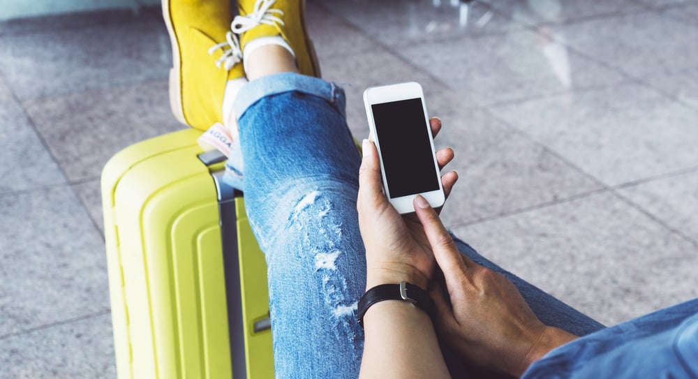 femme assise dans un aeroport avec les pieds appuyes sur sa valise en utilisant son telephone portable